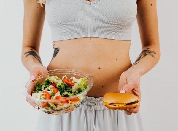 A woman in a sports bra holding a salad and cheeseburger, representing healthy vs junk food choices.