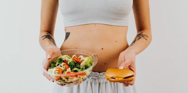 A woman in a sports bra holding a salad and cheeseburger, representing healthy vs junk food choices.